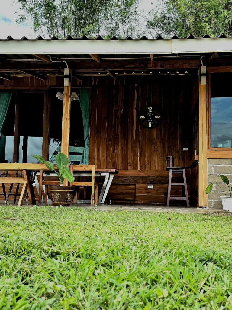 A teak porch with curtains and a clock, framed by tall grass at twilight.